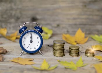 Alarm clock and money coins on wooden rustic old table with autumn leaves. Time is money autumn style comcept. Discount time concept. Time for fall autumn savings. Time to make money.